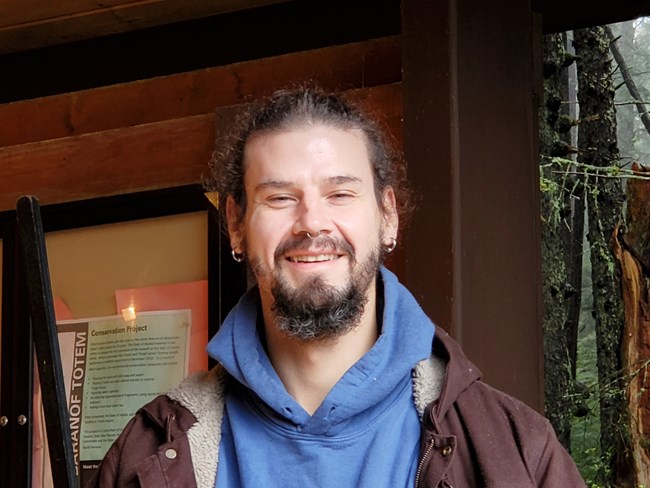 A smiling man. The man has brown hair and has a mustache and beard. He is wearing a blue sweatshirt and brown jacket. The man has a silver nose ring and earrings.