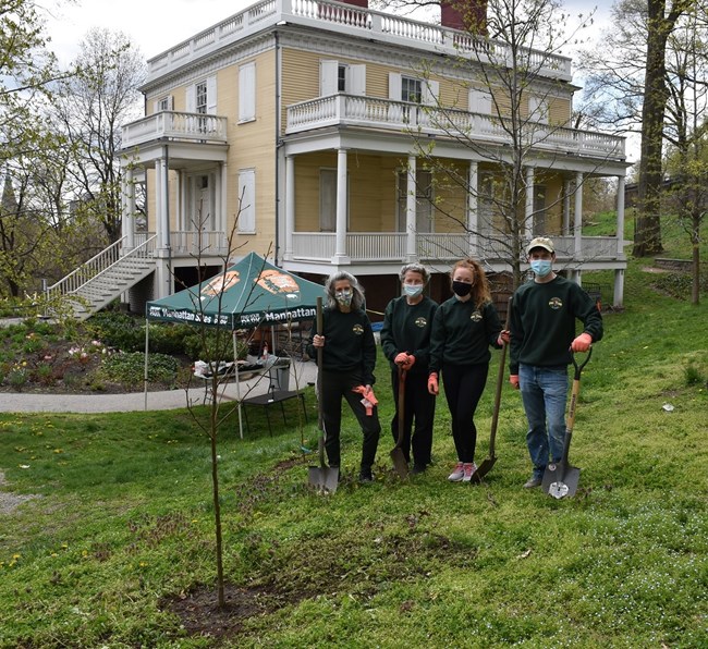 A group of four people stand around a tree sapling, holding shovels.