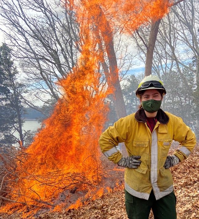 Ignitions at Salt Pond Visitor Center's burn pile on March 16th 2021. Woman wearing yellow protective equipment stands next to a fire.