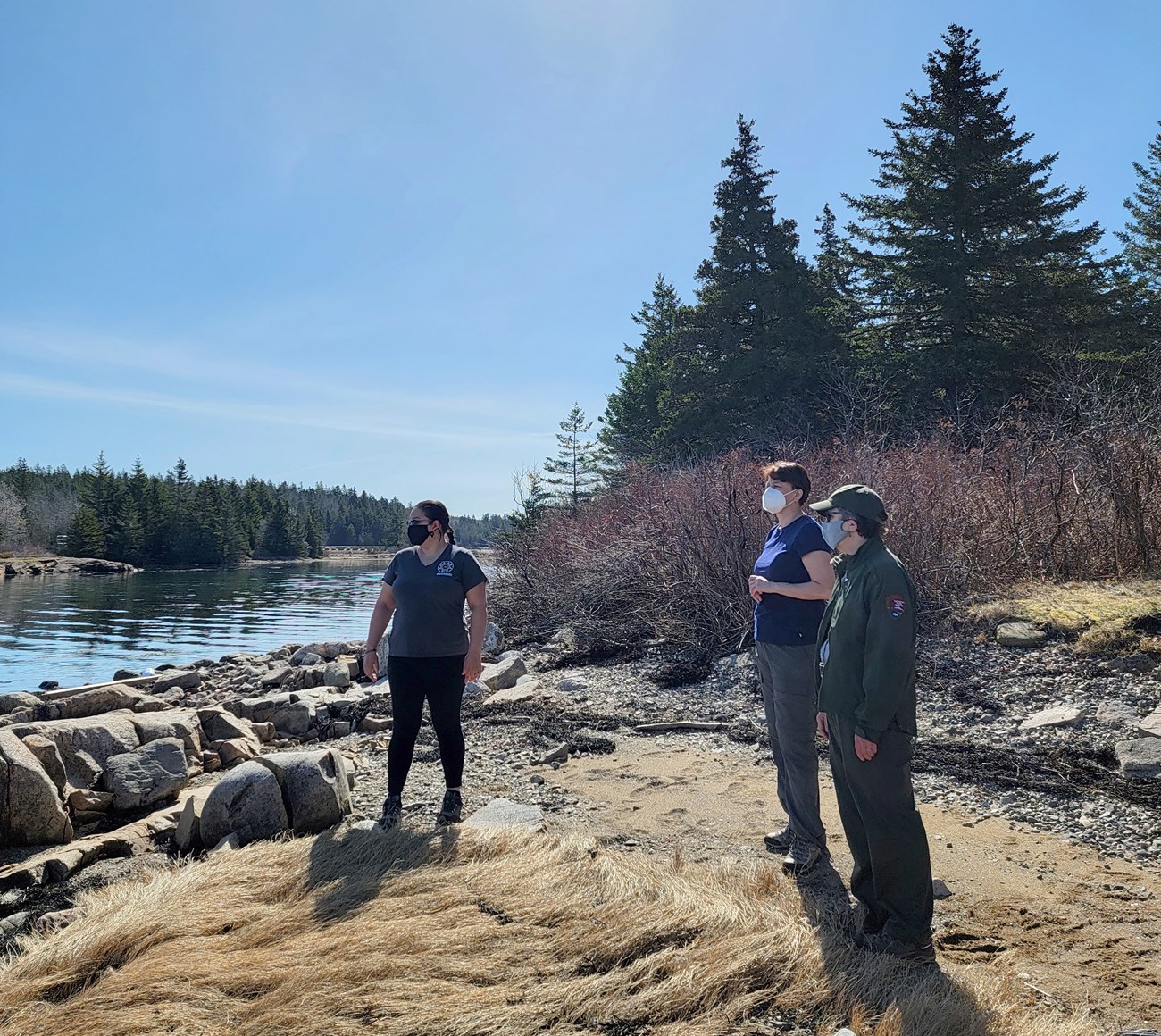 Two women and one man with face masks look at the shoreline of a lake. Evergreen trees are behind them and across the lake. The man is wearing a green Park Service uniform.