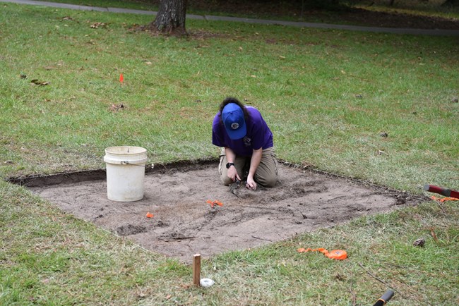 A person sits in a square shallow pit looking intently at the dirt ground around them.
