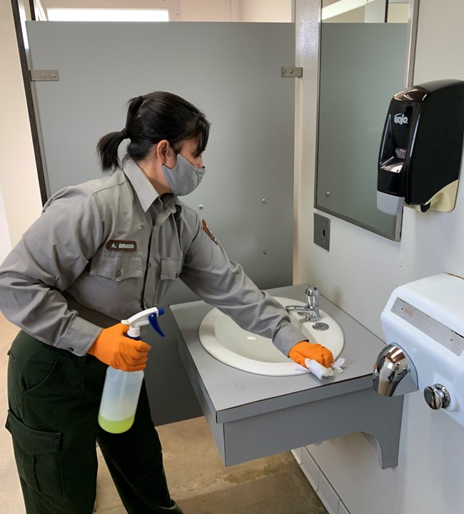 Female uniformed ranger wearing orange gloves holds a spray bottle in one hand and wipes a bathroom sink with the other.