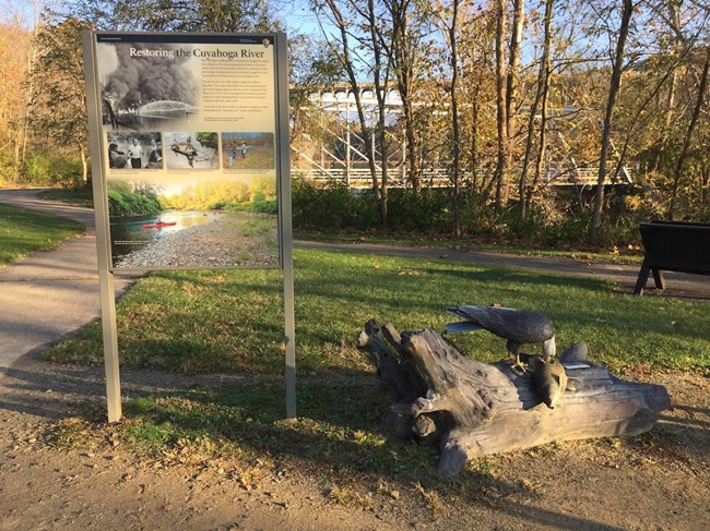 A display board with an eagle statue next to it with grass, trees and a bridge in the background.