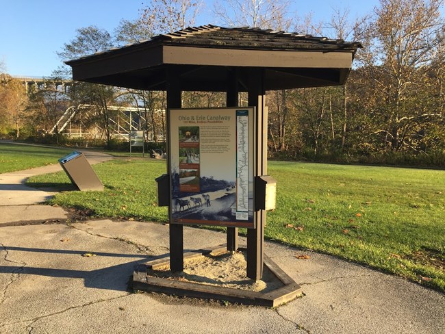 Wooden, three-sided informational kiosk with grass, trees and a bridge behind it.