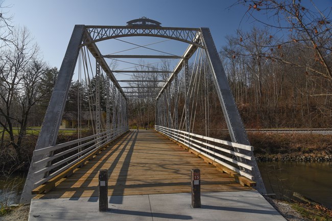 SRB freshly painted View down a white metal truss bridge with a wooden floor spanning a river; railroad tracks and bare trees on the far side.
