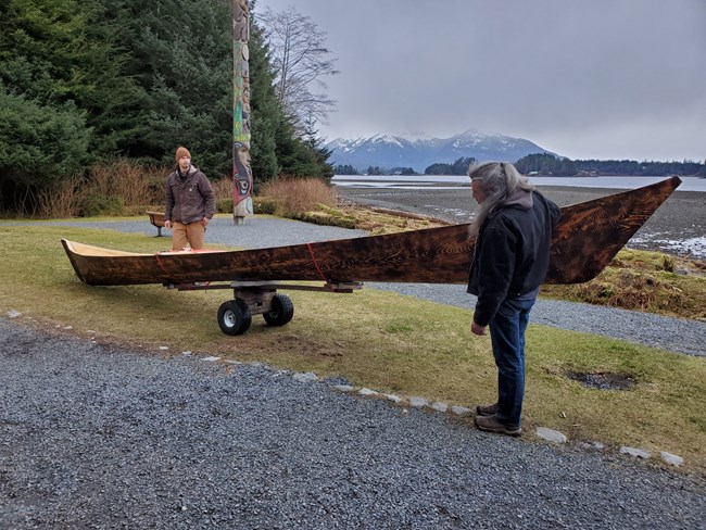 On a patch of grass next to a path, two men begin to move a dugout canoe towards the receded ocean with snowcapped mountains in the distance.