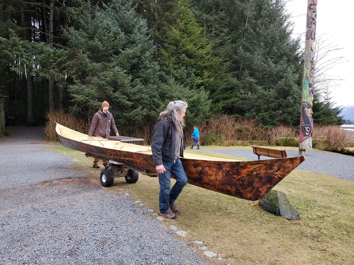 Two men hauling a large wooden canoe on a small trailer alng a gravel path. A forest is in the background.