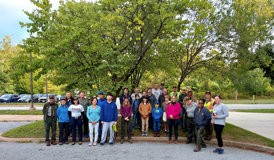 A group of smiling people stand outside, under trees in a parking lot