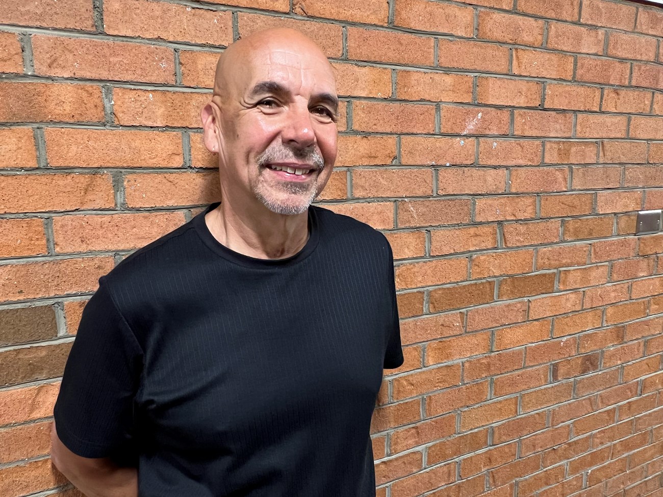 A smiling, bald man with a grey beard stands against a brick wall.