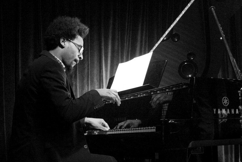 A Black man wearing a blazer and glasses plays a grand piano in front of a stage curtain.