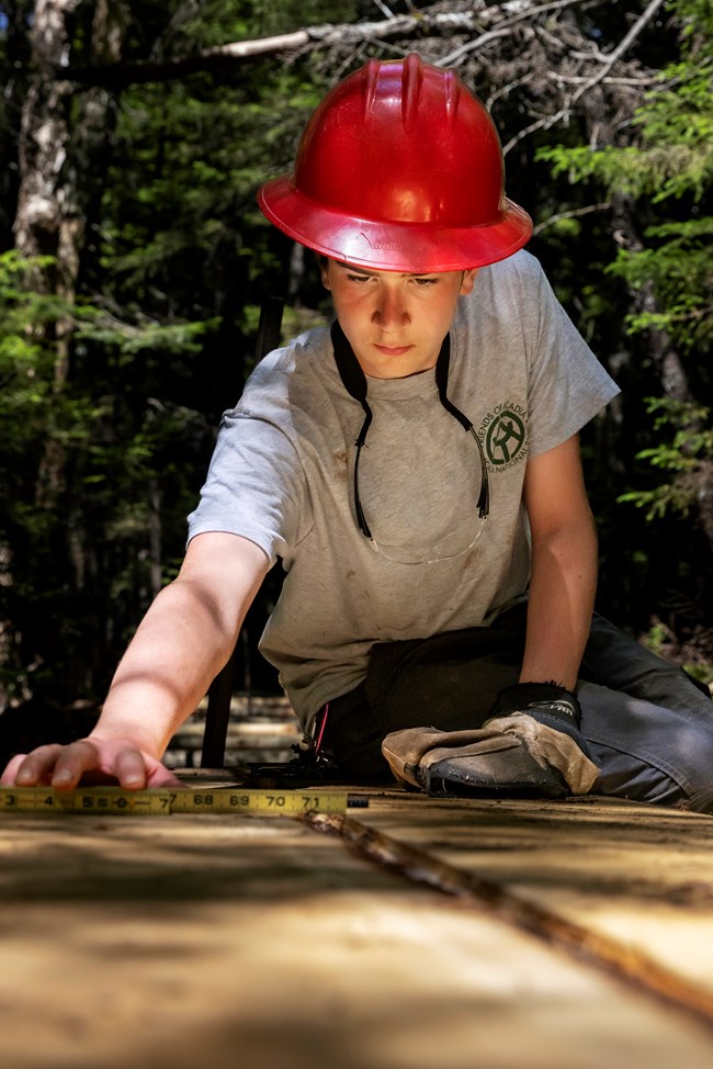 Boy in work uniform and red hard hat uses a measuring tape on a boardwalk