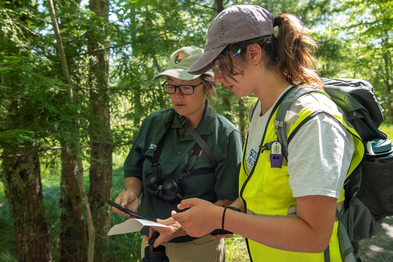 Two women in uniform in an outdoor setting have a conversation while holding electronic devices.