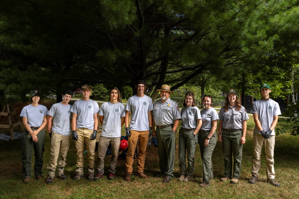 Group photo of nine young people in work clothes and uniforms with a man a center wearing a ranger flat hat and NPS uniform