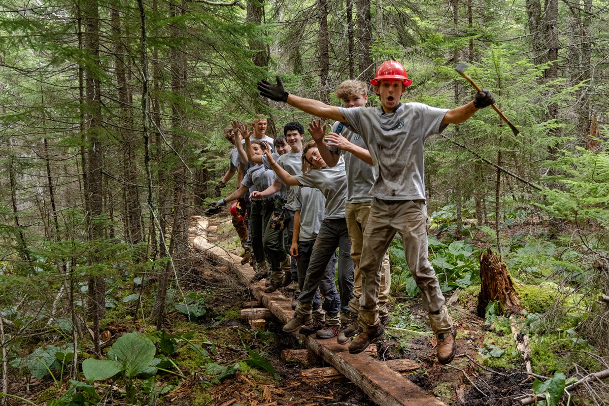 In a dense wooded area, eight young people in work clothes and red hard hats jump and exclaim for the camera