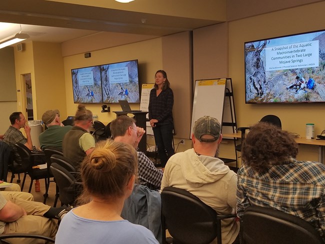 View of people seated in a large room listening to a woman standing up front giving a presentation that shows up on a screen behind her.
