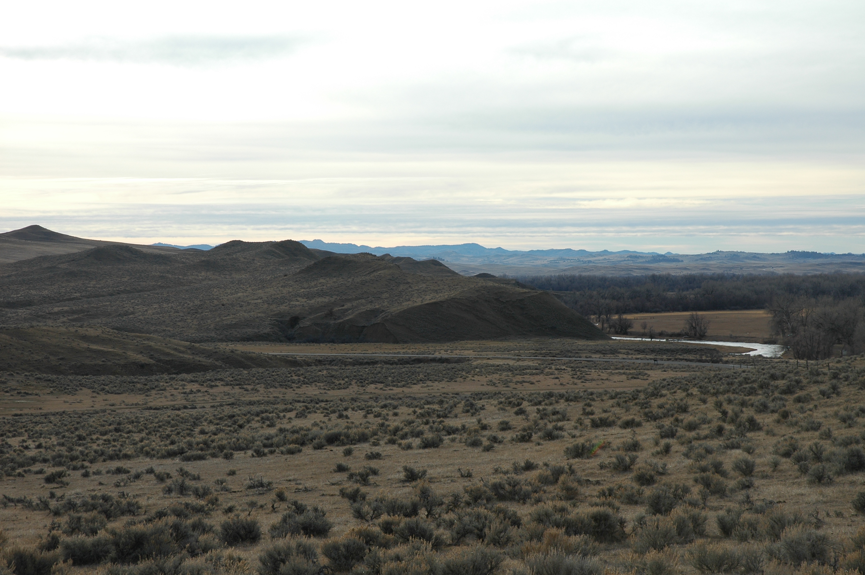 Little Big Horn Battlefield A large open prairie near several low gray hills and a small creek in the left-hand side of the image foreground. A distant chain of mountains are visible in the background. A small forest can be seen in between.