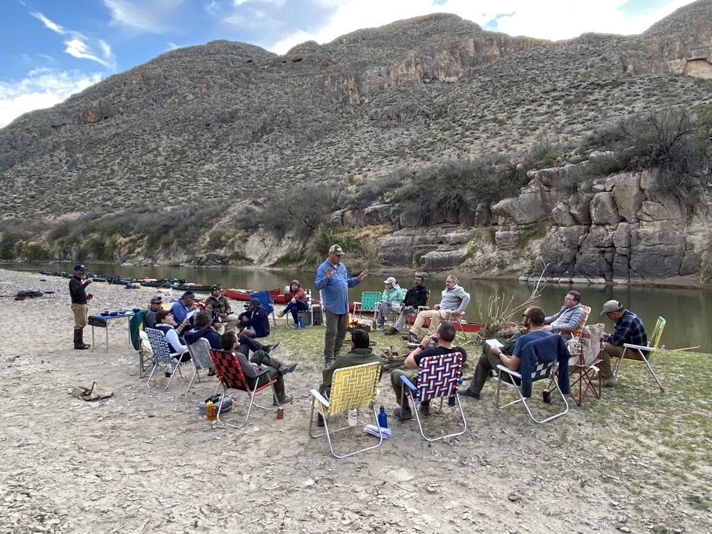 Big Bend and Mexican park staff gather around a campfire in the evening on a rocky river shore.
