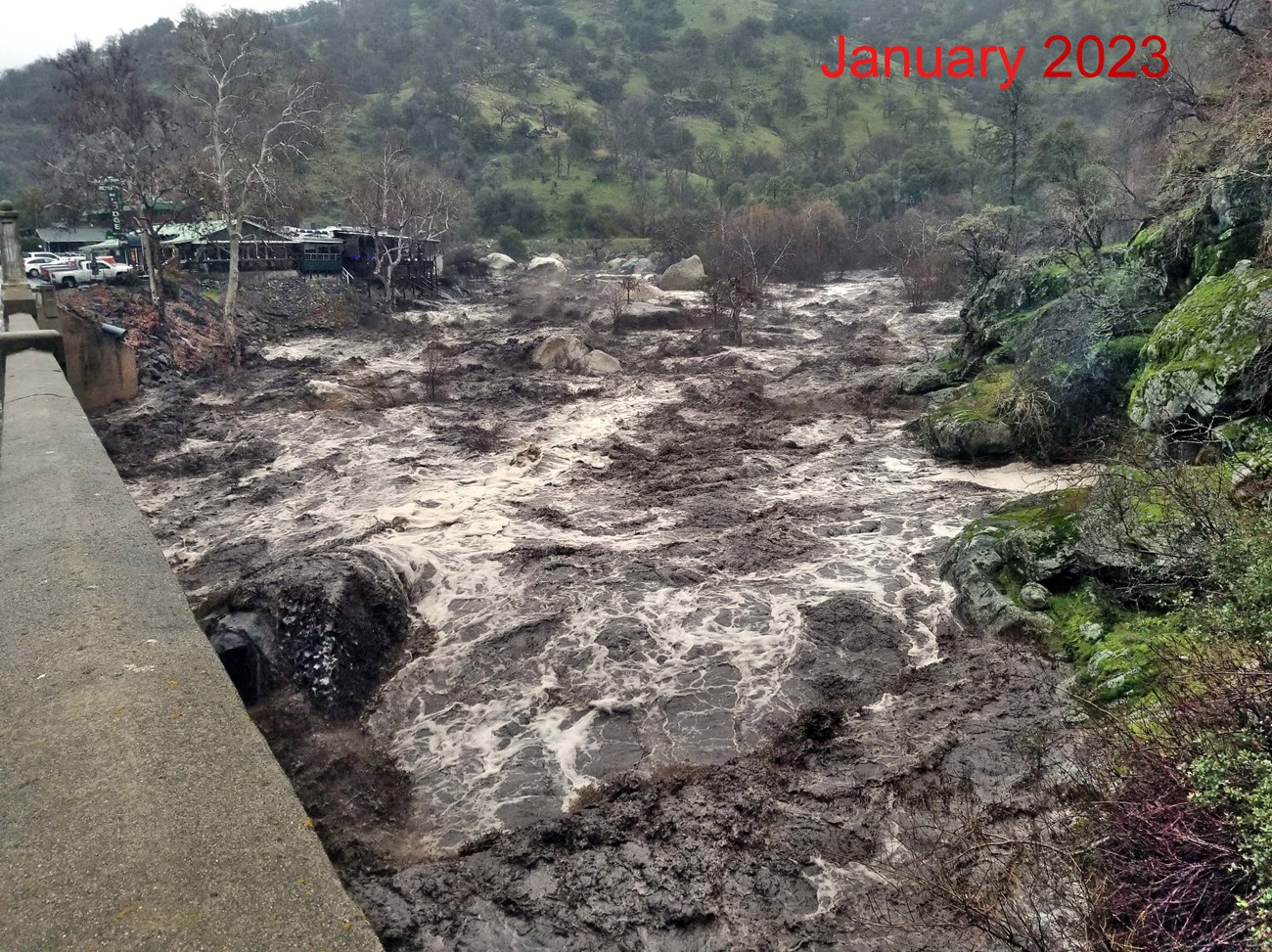 View from bridge of dark, muddy water of river flowing high enough to cover most large rocks in river channel.