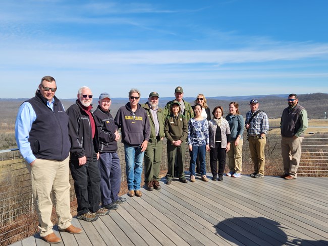 13 individuals smiling for a group picture on the Summit Bechtel Reserve deck. Some are wearing sunglasses, hats, and others are in park uniforms. Trees and blue skies can be seen in the background.