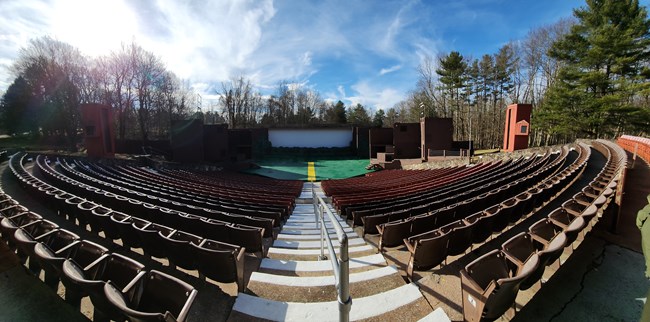 Cliffside Amphitheater A panoramic view of the Cliffside Amphitheater with pullout seats on either side. Stairs with a grey railing appear in the center, which leads to the stage. The stage is marked by a yellow line in the center, green floor, and a white projection screen. In