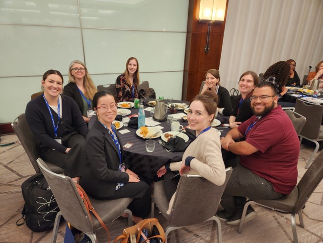 8 individuals sit around a circular table during lunchtime. They are dressed in business casual clothing.