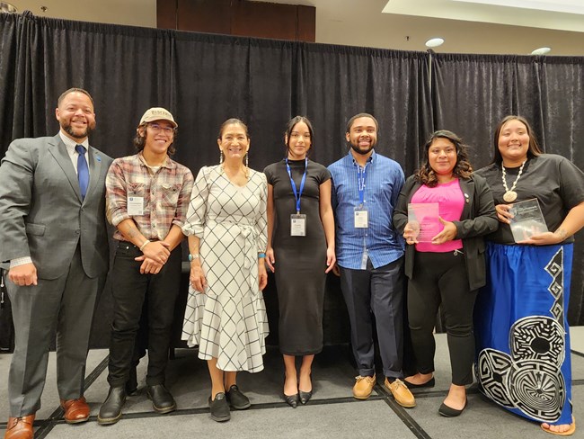 Group photo of 2023 Corpsmember of the Year Awardees 7 individuals from different backgrounds smiling for a group photo. The clothing is a mix of business professional and business casual. The two women on the far right are holding up rectangular glass awards.