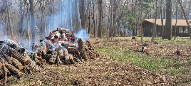 Piles burning behind Furnessville Rd Dorm 2 large piles of logs, branches and sticks smolder with a bron 1 story house and leafless trees in the background