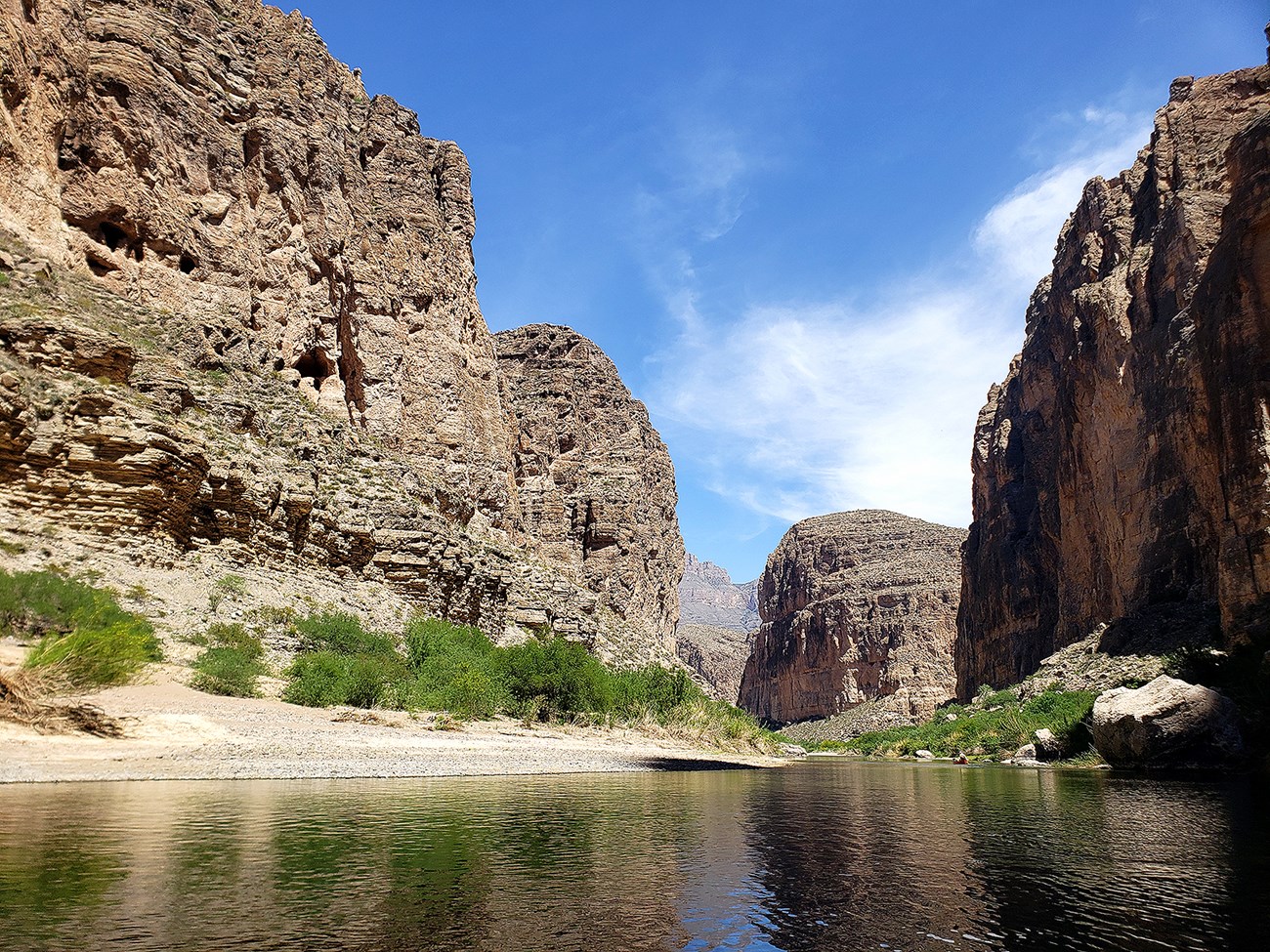 Gray canyon walls tower over a river with the blue sky reflected in it.