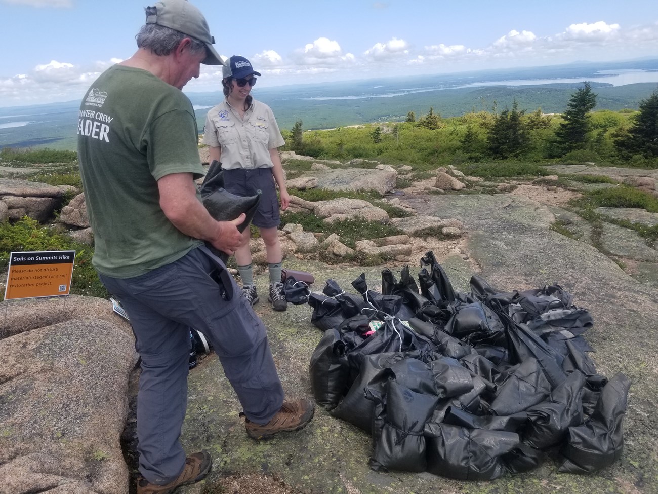 Save our Summits event Two park volunteers in outdoor clothing carry black plastic sandbags on a granite summit,