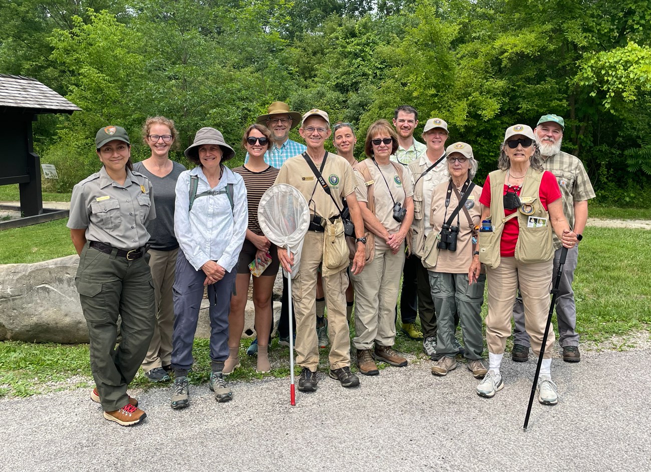 Thirteen people stand smiling outdoors; one is a uniformed ranger and several others wear khaki shirts and vests, holding binoculars and nets.