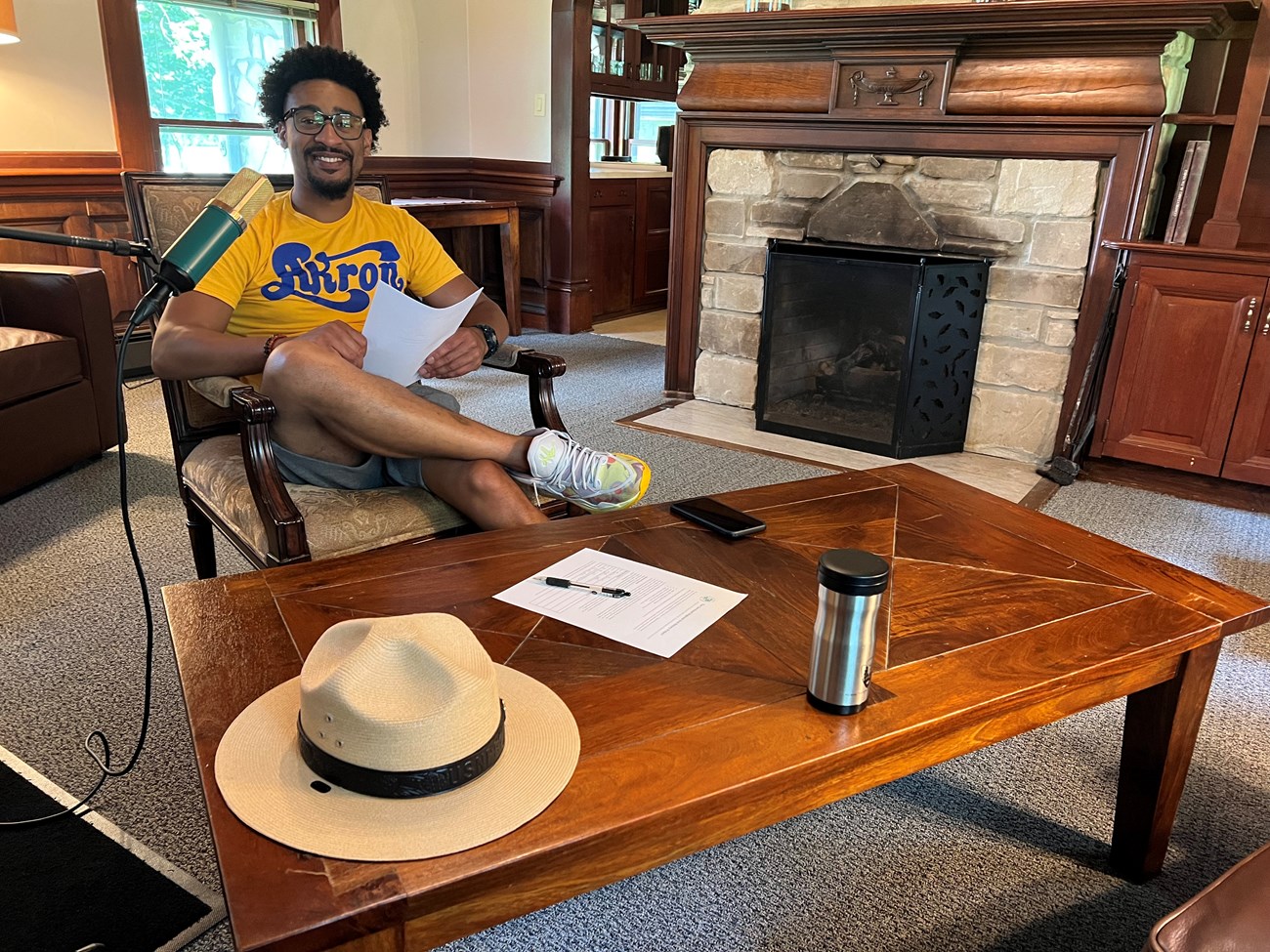 A Black man sits behind a microphone, a stone fireplace to the right. He has a short afro and beard, wears glasses and a yellow and blue Akron shirt.