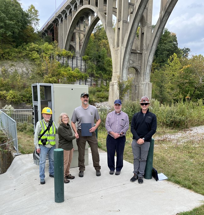 A woman with blond hair and glasses poses with four men, two wearing hard hats, in front of a pump station under an arched bridge.