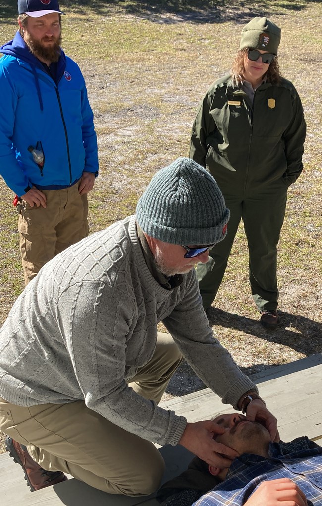 Man holding a another man's face while he is laying on the ground. Two people look on in the background.