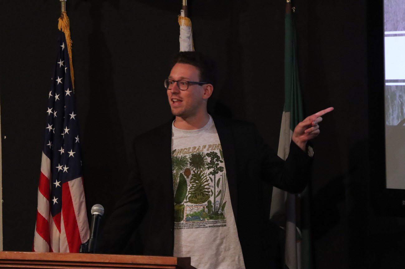 Dr. Ben Morris a man at a podium speaking to an audience while pointing to a screen.