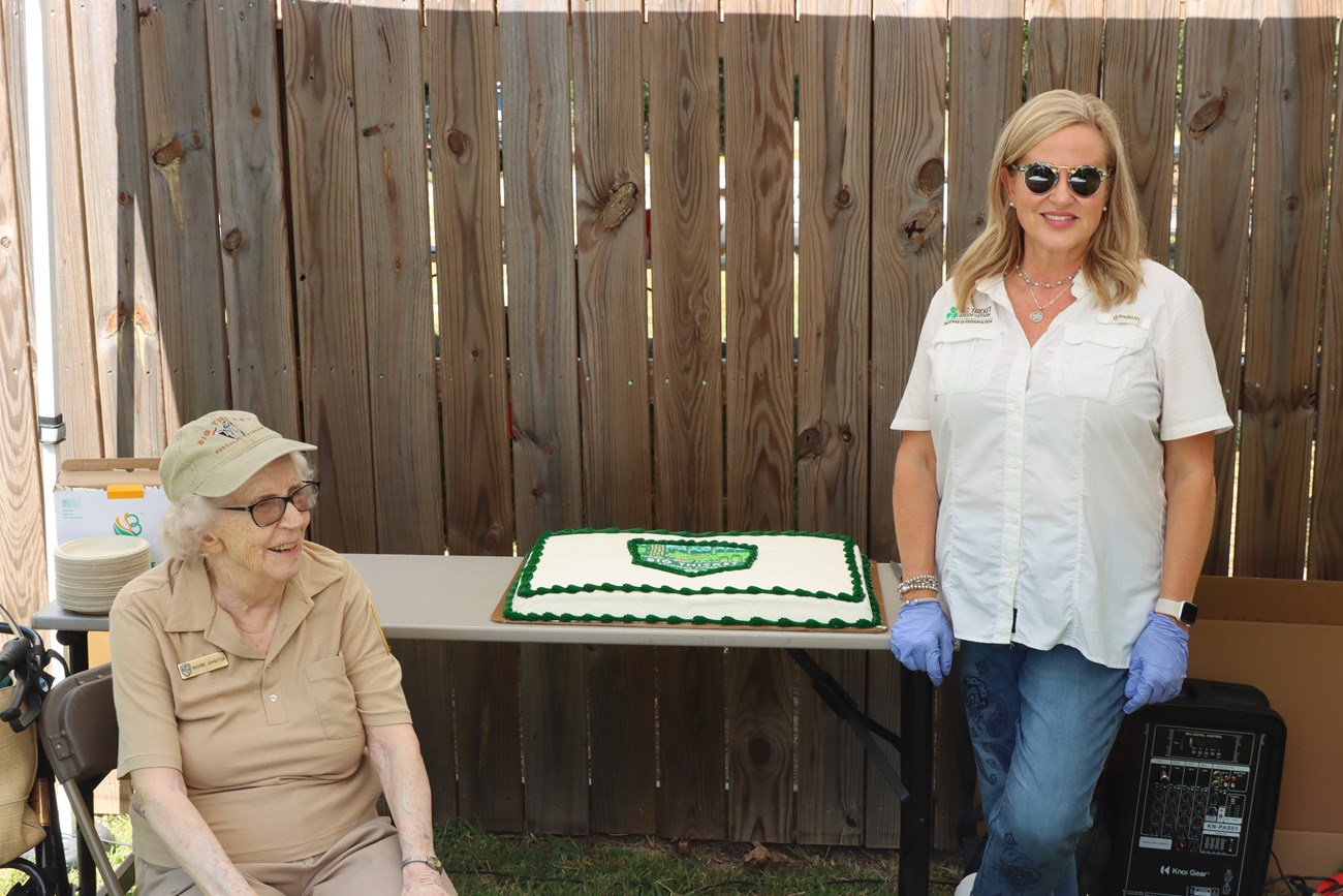 Maxine and Diane 1 woman sitting and another woman standing, in front of a white cake sitting on a table in front of a wooden fence.