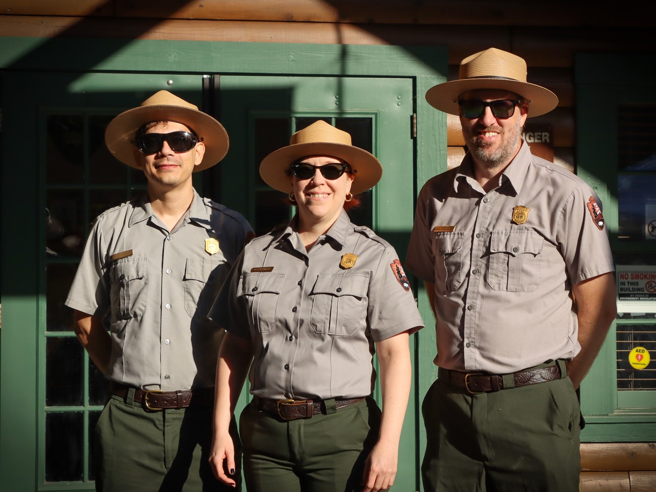 Park rangers 3 park rangers wearing dark sunglasses, flat hats, gray shirts, and green pants, posing outside the visitor center.
