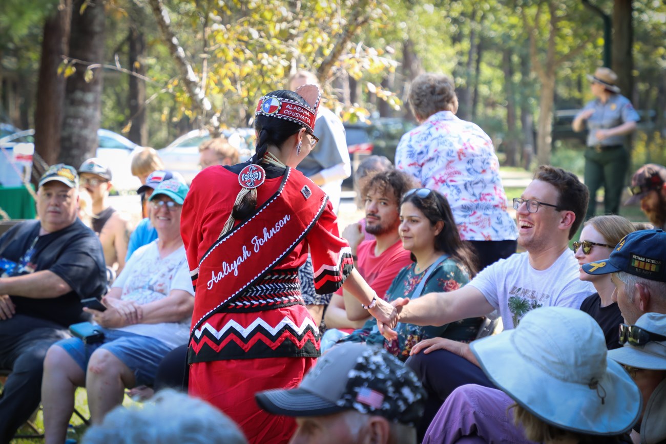Alabama-Coushatta performance a woman in bright red regalia shaking hands with people in the audience at an outdoor event.