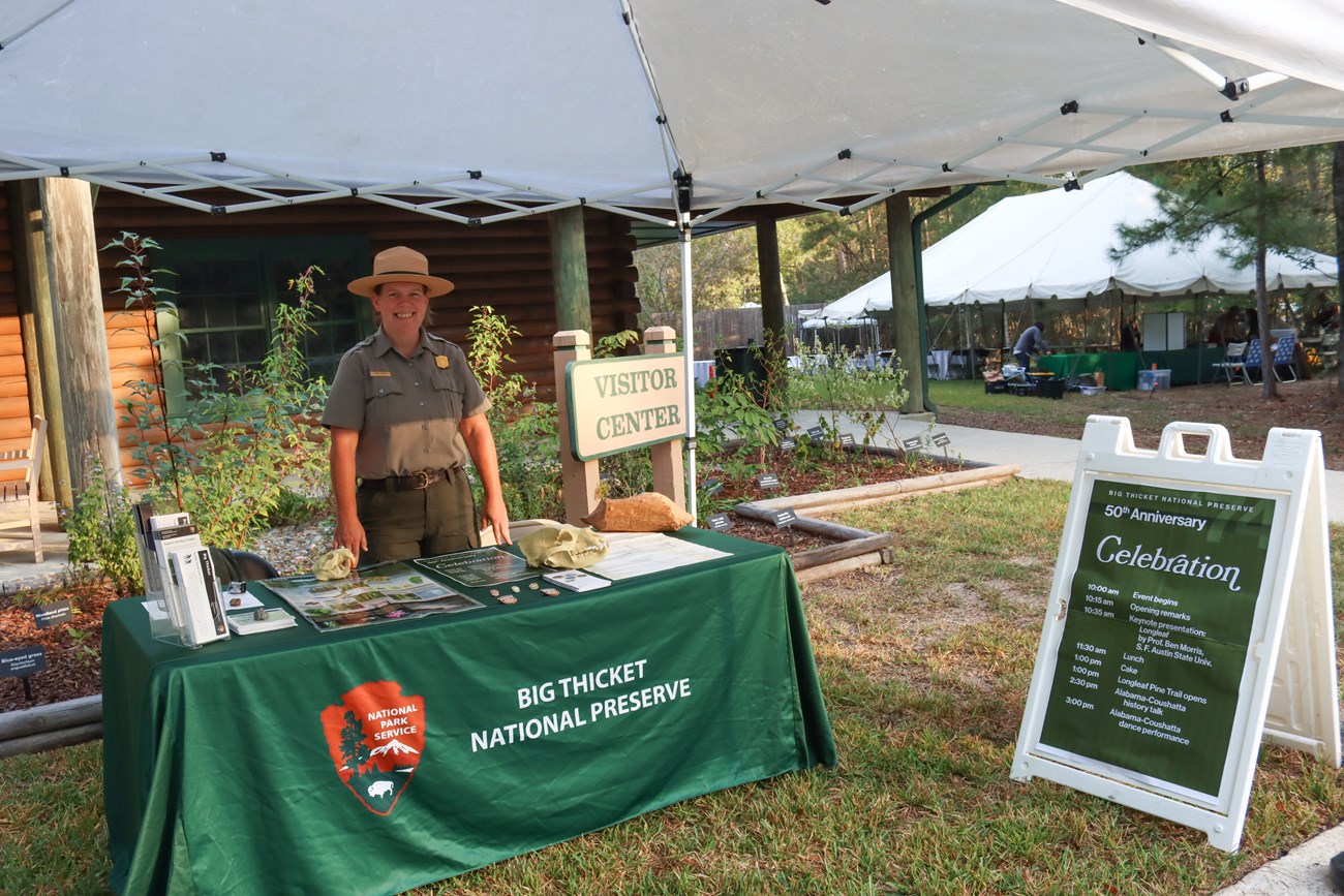 welcome table a park ranger stands at a green table with props on it, next to a sign that says visitor center. She stands below a white canopy.