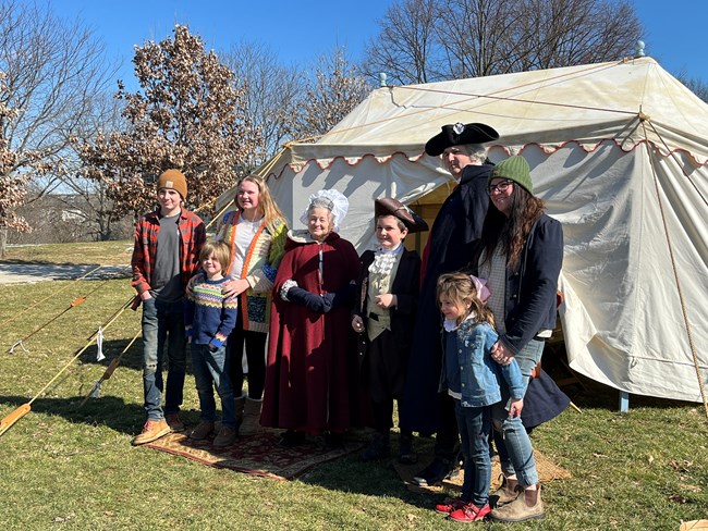 Washington's Birthday 2024 A family of six pose with a man and a woman wearing 18th century clothing, reenactors portraying george and martha washington in front of a cloth tent.