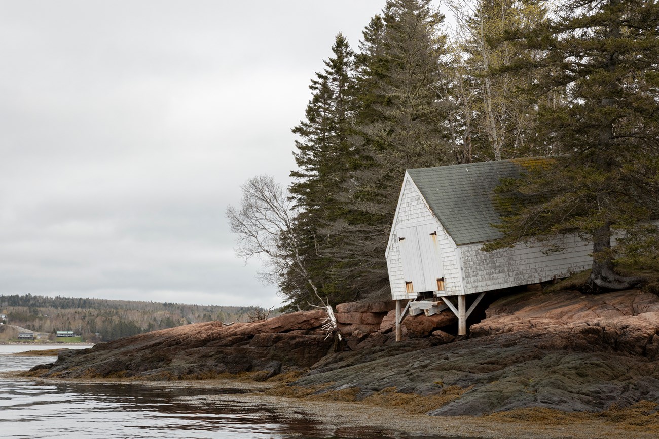 Saint Croix Island International Historic Site after winter storms A white, wood building leans toward water out of a forest of pine trees