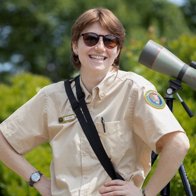 SCA Raptor Intern Sara Smiling SCA Raptor Intern Sara poses outdoors next to a tripod-mounted spotting scope pointed at a peregrine falcon fledgling.
