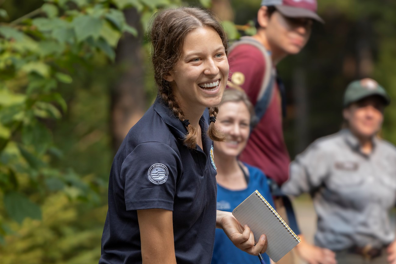 A smiling woman talks to a crowd of volunteers while holding a notepad and pen.