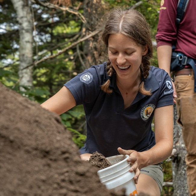 Claire filling bags of soil for volunteers to carry. A woman scooping dirt from a large pile into a white bucket.