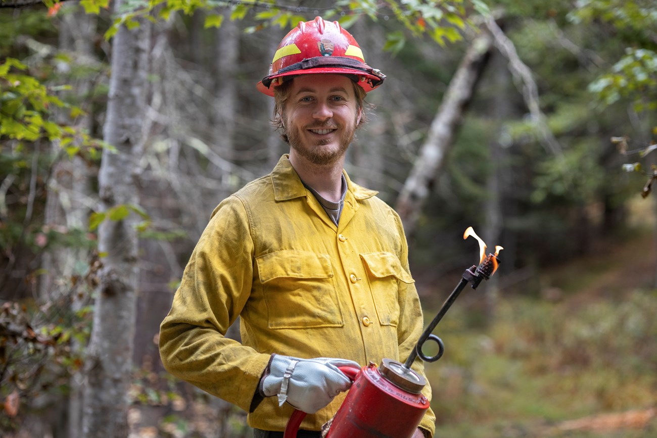 Aidan Gordon, Wildland Fire Apprentice, holds a flaming drip torch in a forested environment. Aidan Gordon, Wildland Fire Apprentice, holds a flaming drip torch in a forested environment.