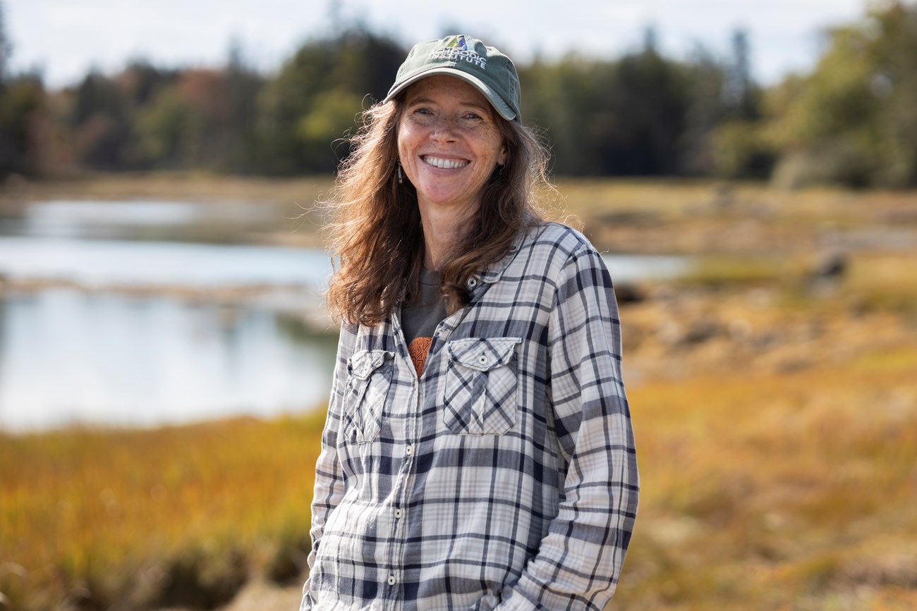 Catherine Schmitt, Science Communication Specialist, smiles while at the Thompson Island Picnic Area. Woman wearing a hat stands in a wetland environment while smiling with the wind blowing her hair to the side.