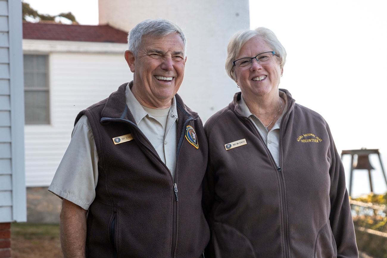 Ray & Mary Ann Schaefer, Volunteer Rangers, smile in front of the Bass Harbor Head Lighthouse. A husband and wife wearing volunteer uniforms smile in front of a lighthouse.