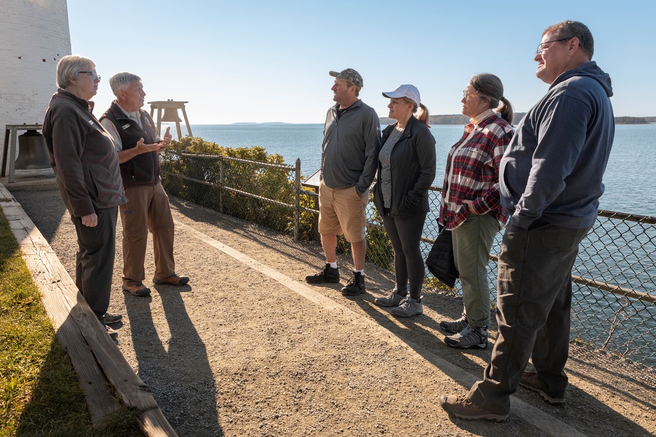 Ray & Mary Ann talk to four visitors in front of the lighthouse. Married volunteers talk to four visitors in front of a lighthouse.