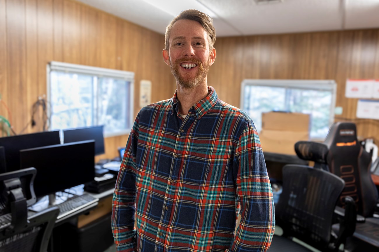 Sean Orr, ---------, stands in his IT office where he works on fixing computer hardware and software. A man wearing a plaid shirt stands in a small office space filled with computers.