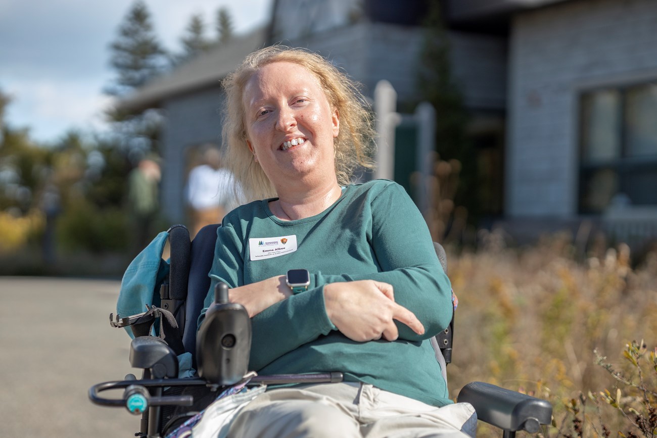 Emma Albee, Science Information Specialist, smiles in front of the Schoodic Institute. Woman in wheelchair smiles in front of Schoodic Institute building.