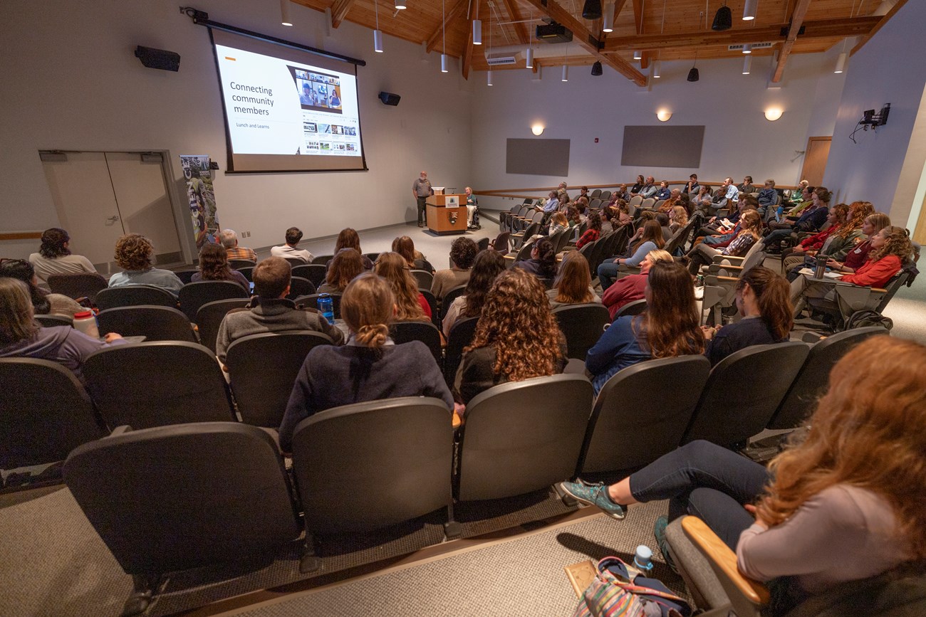 Emma speaks in front of a large crowd in attendance to the Science Symposium. Woman speaks in front of a large crowd in attendance at a conference..